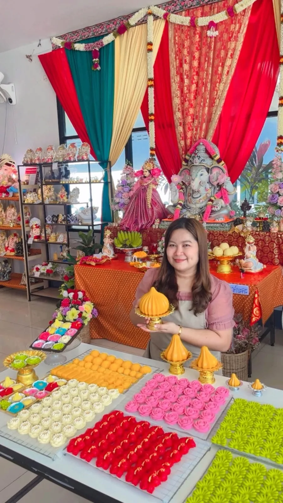 ครูทรายถือขนมโมทกะ ข้างถาดขนมไทยหลากสีที่ปั้นสดในร้าน
